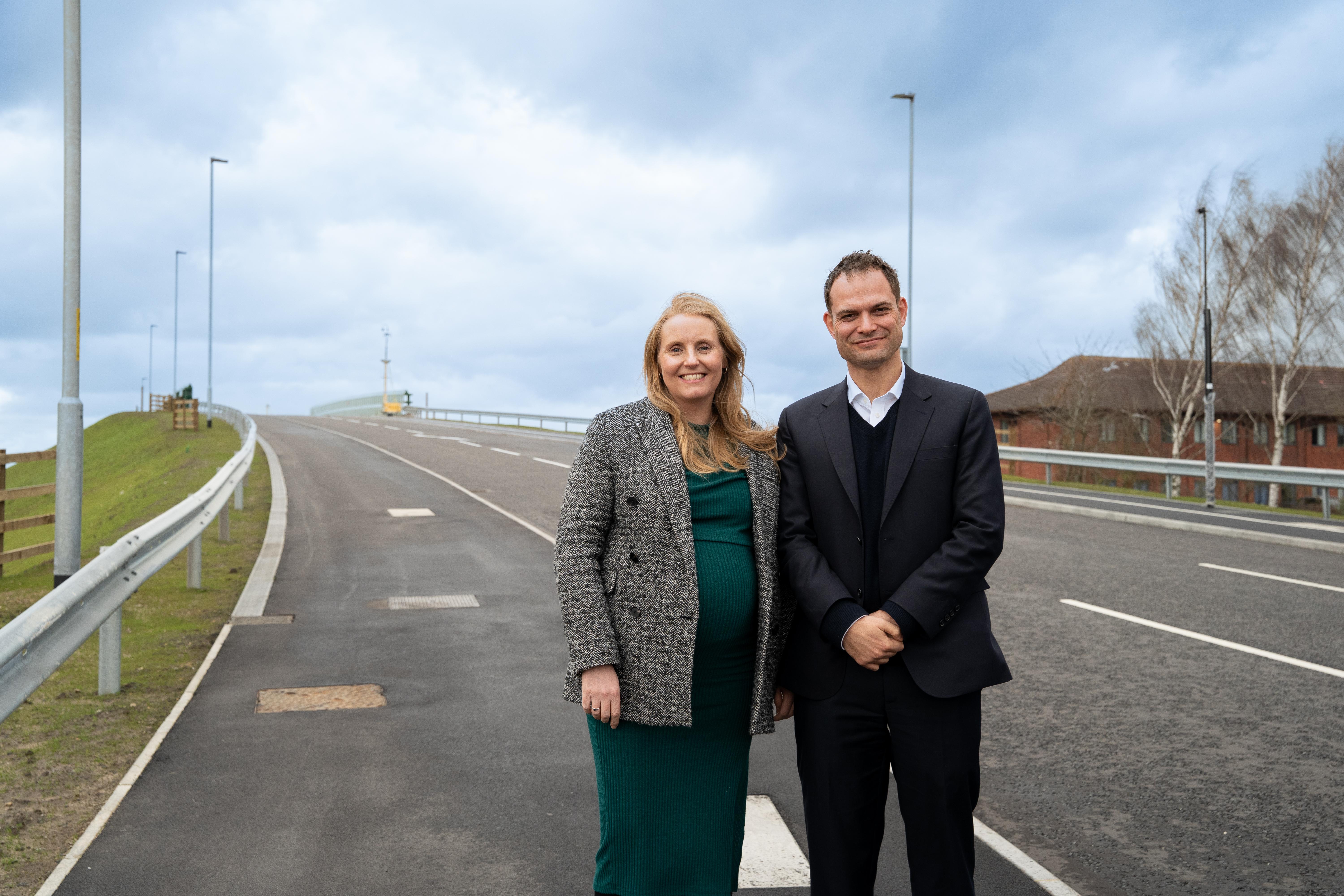 A white woman and man dressed smartly stood together smiling for a photo on a new road bridge that has been built.