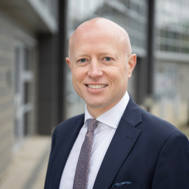 Andrew stands outdoors smiling at the camera in a suit and tie, a building is in the background.